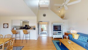 A spacious living area with a dining table, blue sofa, and a TV stand. A kitchen with modern appliances is visible in the background.