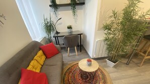Uma sala de estar moderna com um sofá cinzento, almofadas vermelhas e amarelas, uma mesa de centro redonda e um tapete estampado. Há uma mesa de jantar com cadeiras e uma planta em vaso ao fundo.