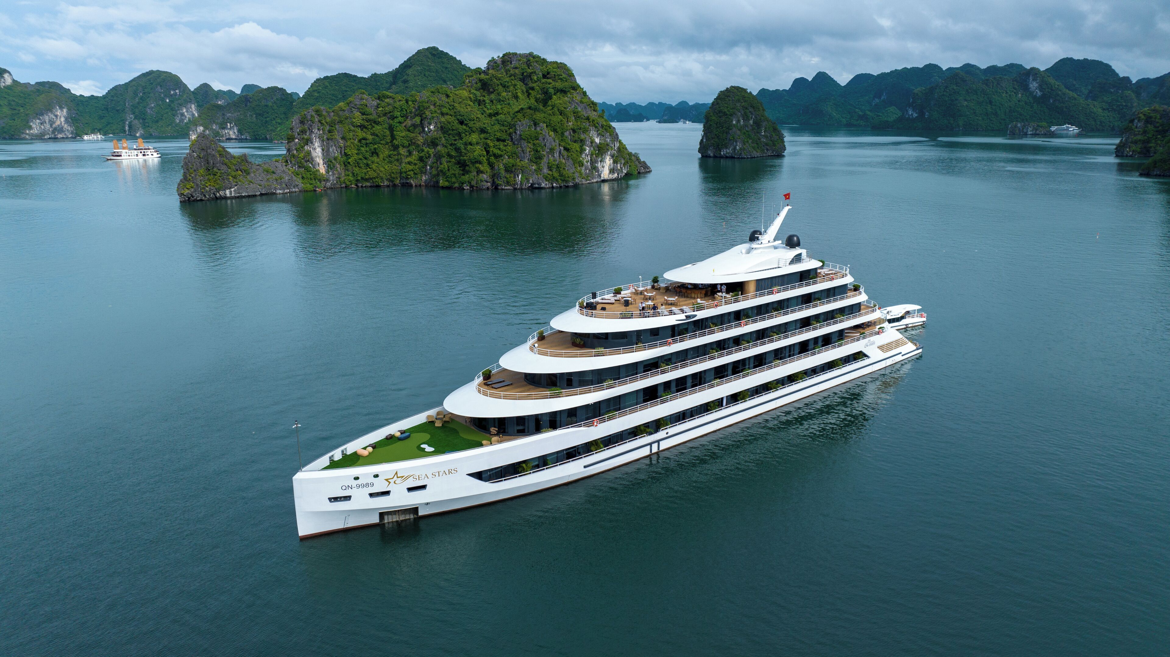 A large, multi-deck cruise ship with outdoor seating areas and a helipad, sailing in a bay with lush green islands in the background.