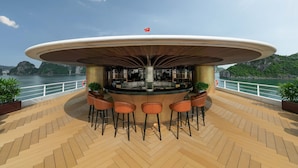 Un espace bar doté d’un toit incurvé, de tabourets de bar et de plantes en pot, situé sur une terrasse en bois offrant une vue sur la mer et les montagnes.