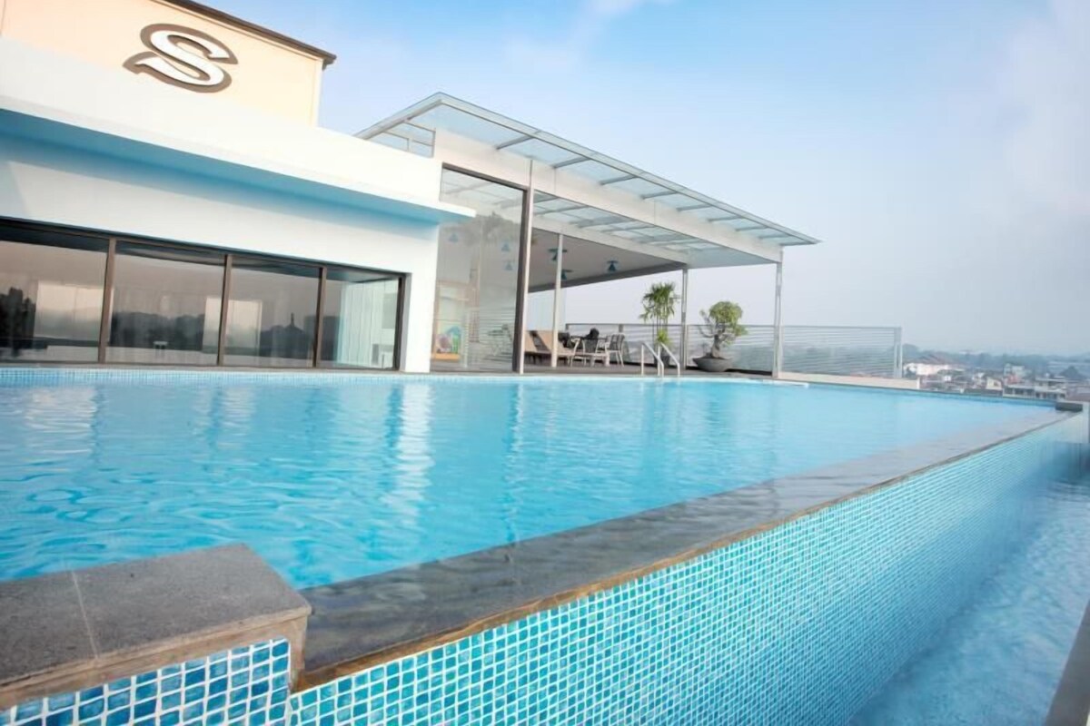A modern hotel with a rooftop infinity pool, a covered outdoor seating area, and a clear view of the city skyline.