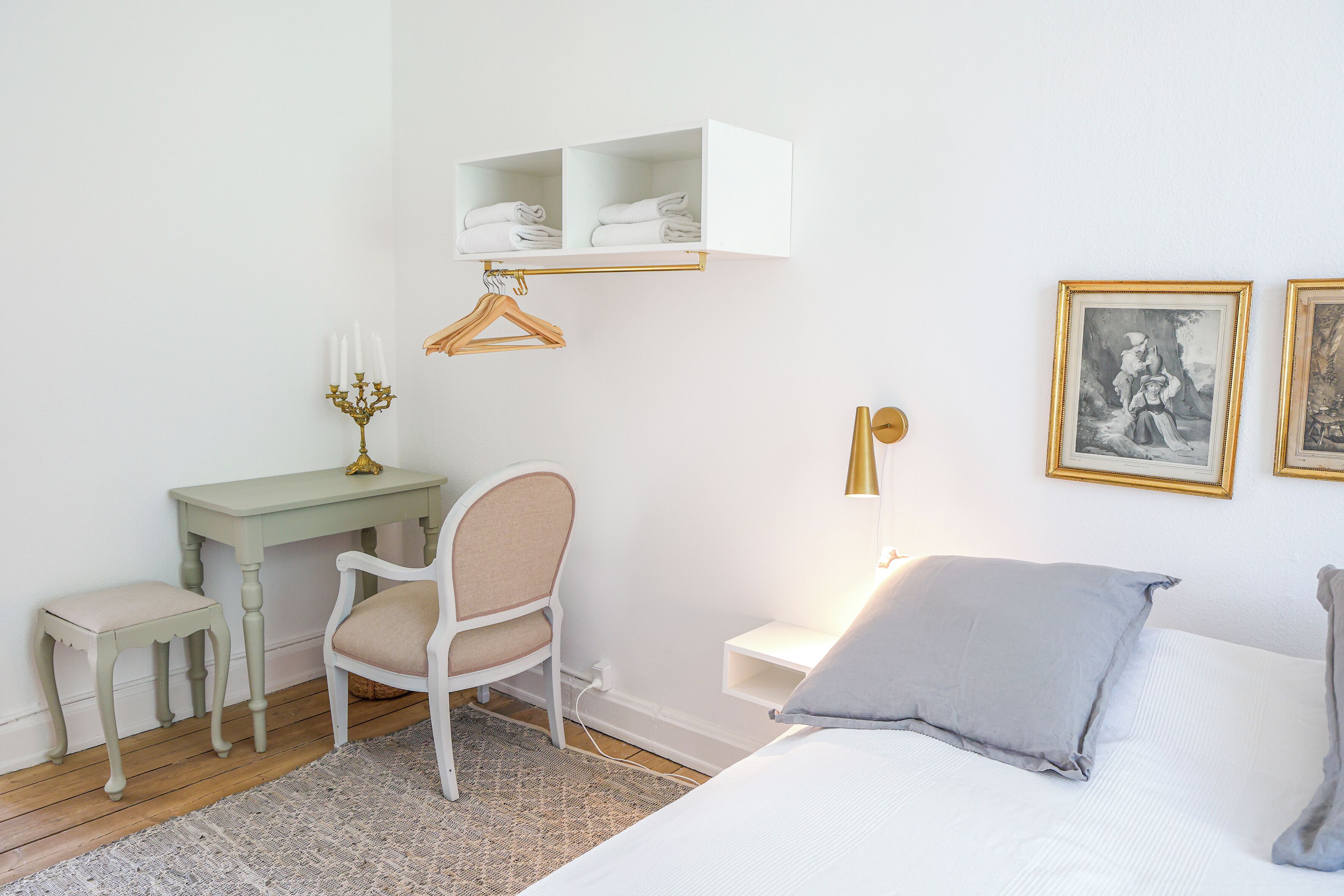 A bedroom with a bed, a desk, a chair, a small table, a hanging shelf, and two wall-mounted lamps.