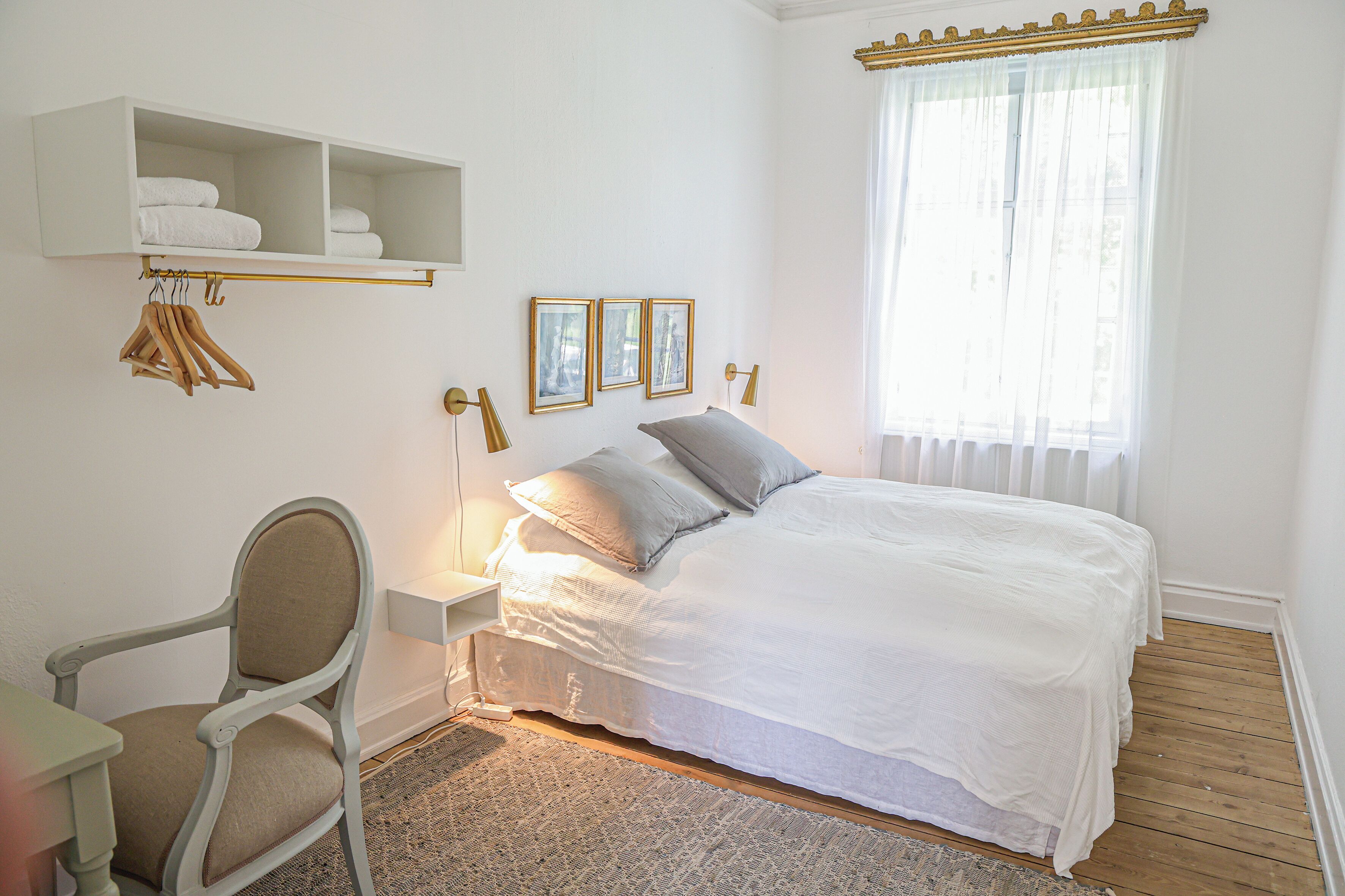 Een slaapkamer met een bed, een stoel, een plank met handdoeken en ingelijste foto’s aan de muur.