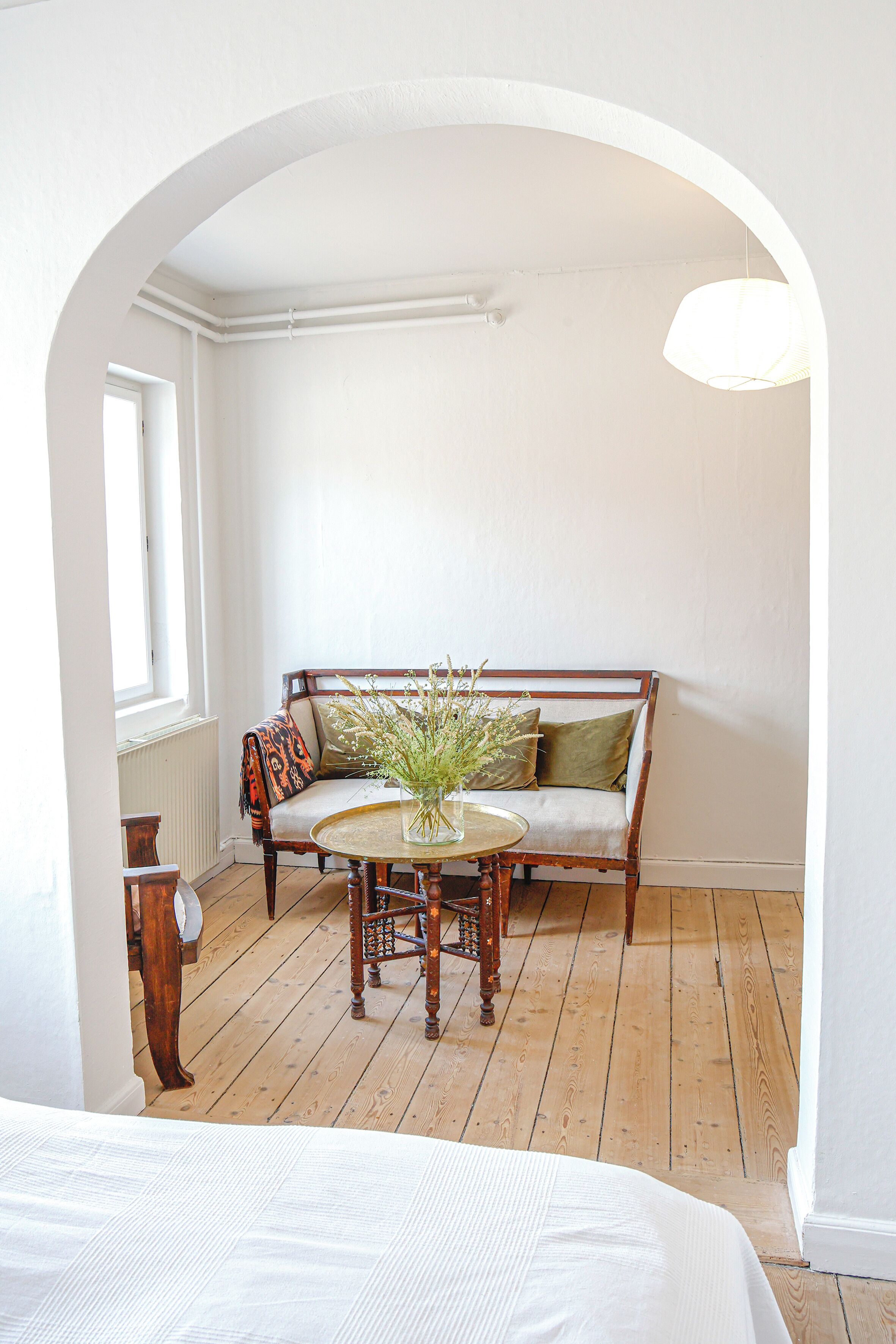 Une pièce avec un plancher en bois, un mur blanc, un banc en bois agrémenté de coussins, une petite table ronde sur laquelle est posé un vase de fleurs, et un luminaire suspendu.