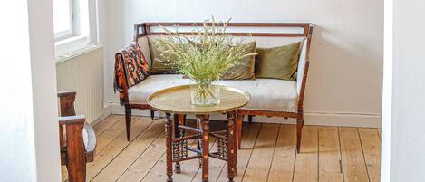 Une pièce avec un plancher en bois, un mur blanc, un banc en bois agrémenté de coussins, une petite table ronde sur laquelle est posé un vase de fleurs, et un luminaire suspendu.