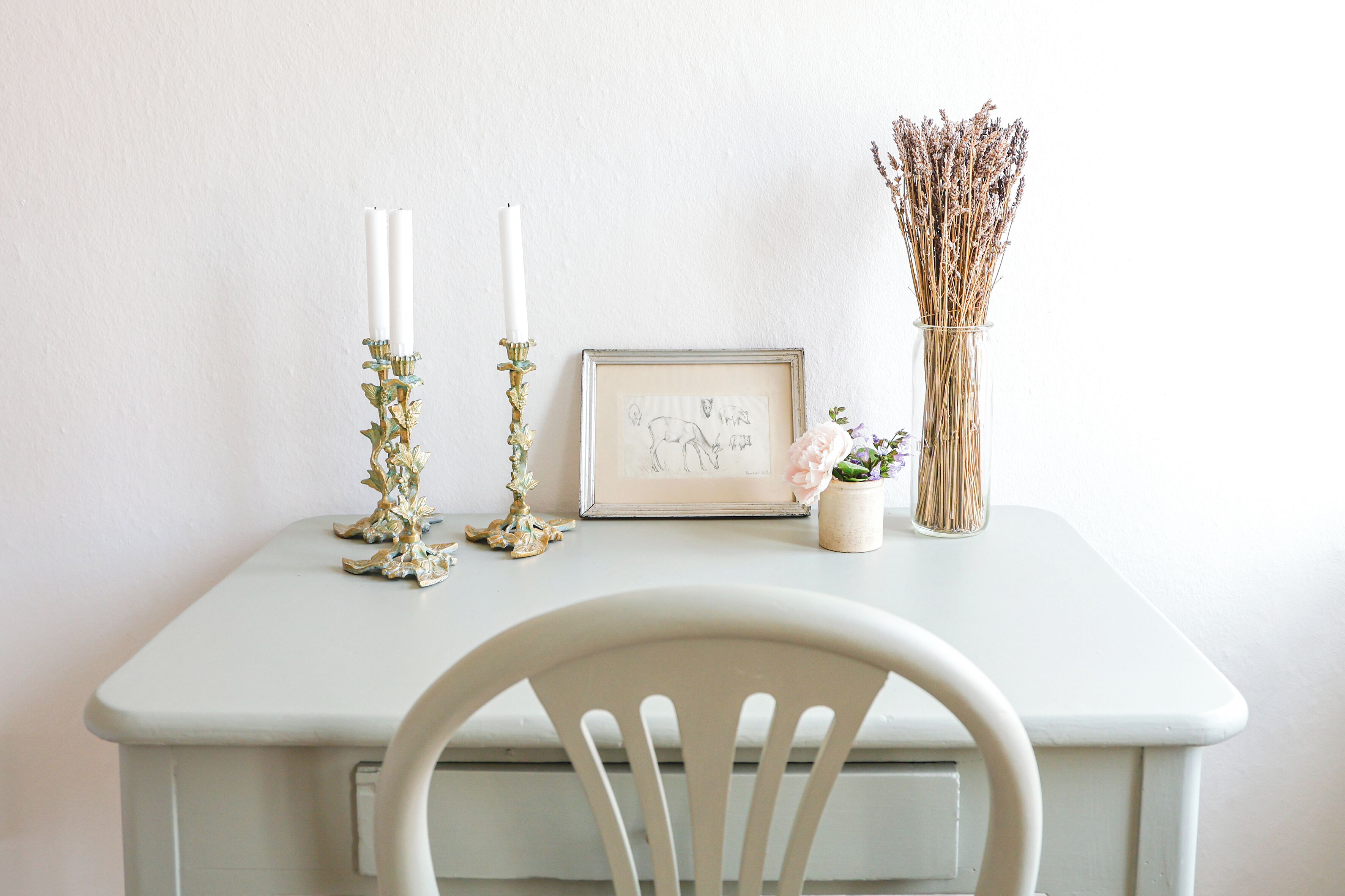 Una mesa blanca con un cuadro enmarco, dos velas blancas, un florero con flores secas y una pequeña planta en maceta.