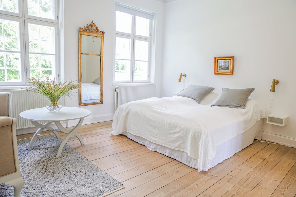 A bedroom with a bed, a round table with a vase, a mirror, and a window.
