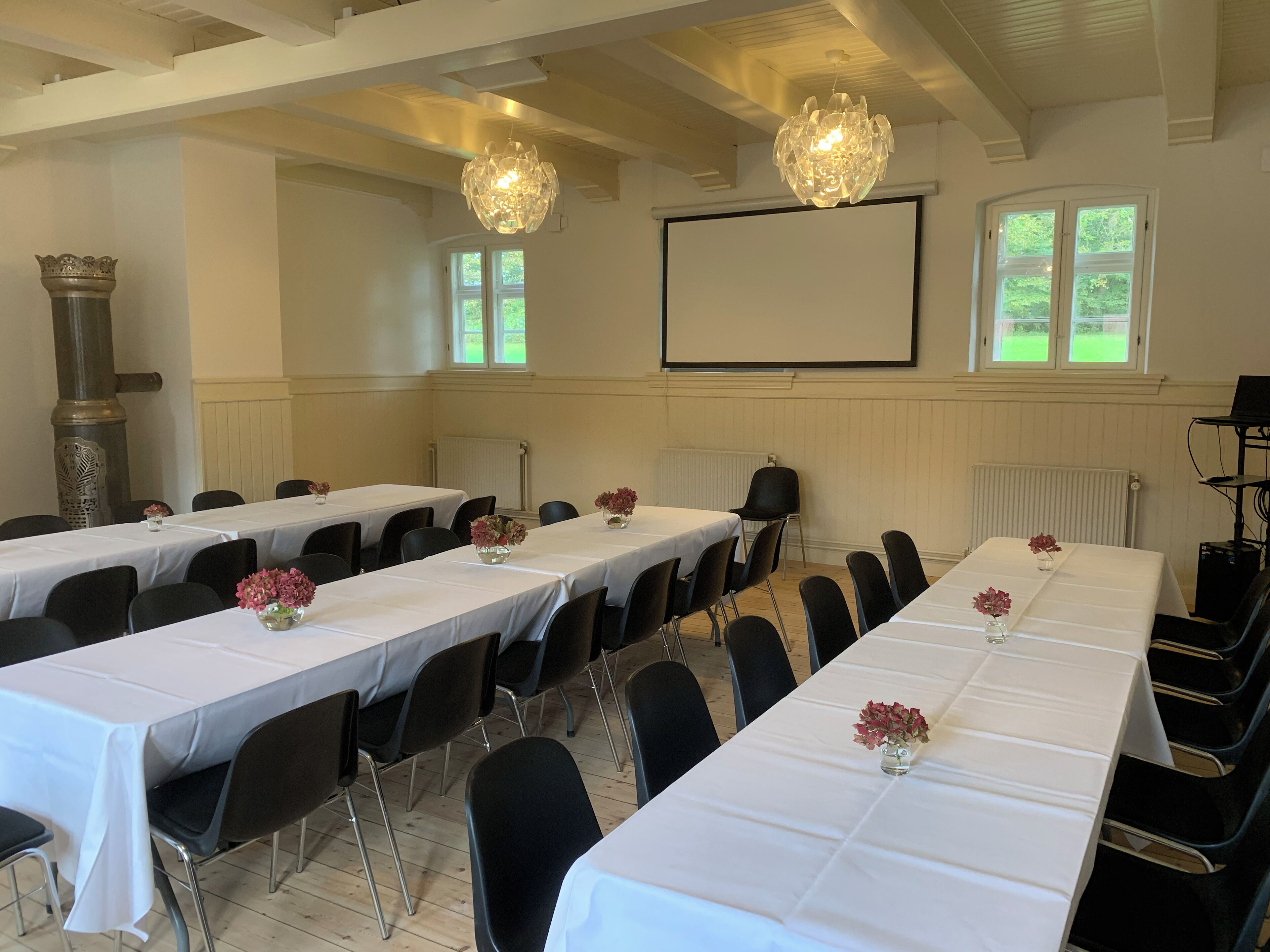 Una sala de conferències amb taules llargues, cadires negres, teles de taula blanques, centrals florals i una pantalla de projector, així com un gran candelabrum decoratiu.