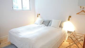 Una cama bien hecha con sábanas blancas y una alfombra azul, una mesita de noche con una lámpara, y un perchero de madera en la pared.