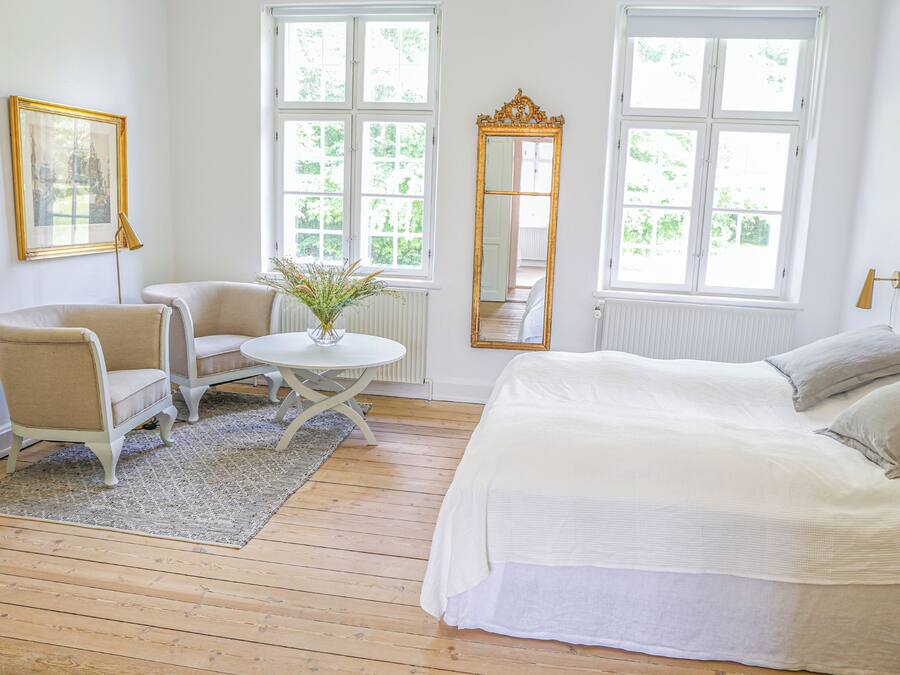 Un dormitorio con una cama, dos sillas, una mesita, un espejo y ventanas amplias.