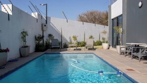 Una piscina rectangular con agua azul cristalina, rodeada por un solárium con baldosas. Se observan sillones de descanso y una mesa a la derecha, y plantas en macetas a la izquierda.