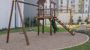 Un parque infantil con una estructura de madera para escalar, un tobogán y columpios, rodeado de grava y césped, con edificios residenciales al fondo.