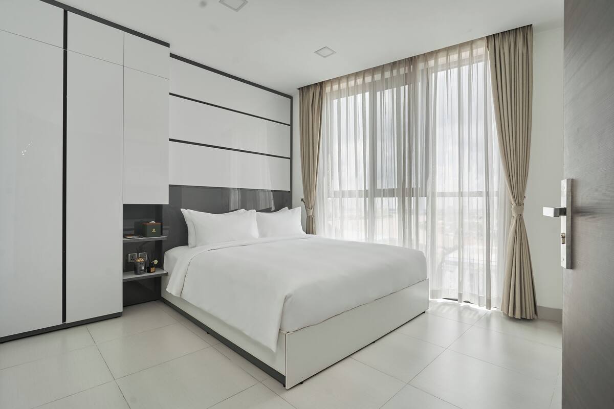 A modern bedroom with a large bed, built-in wardrobe, and a view of the city through sheer curtains.