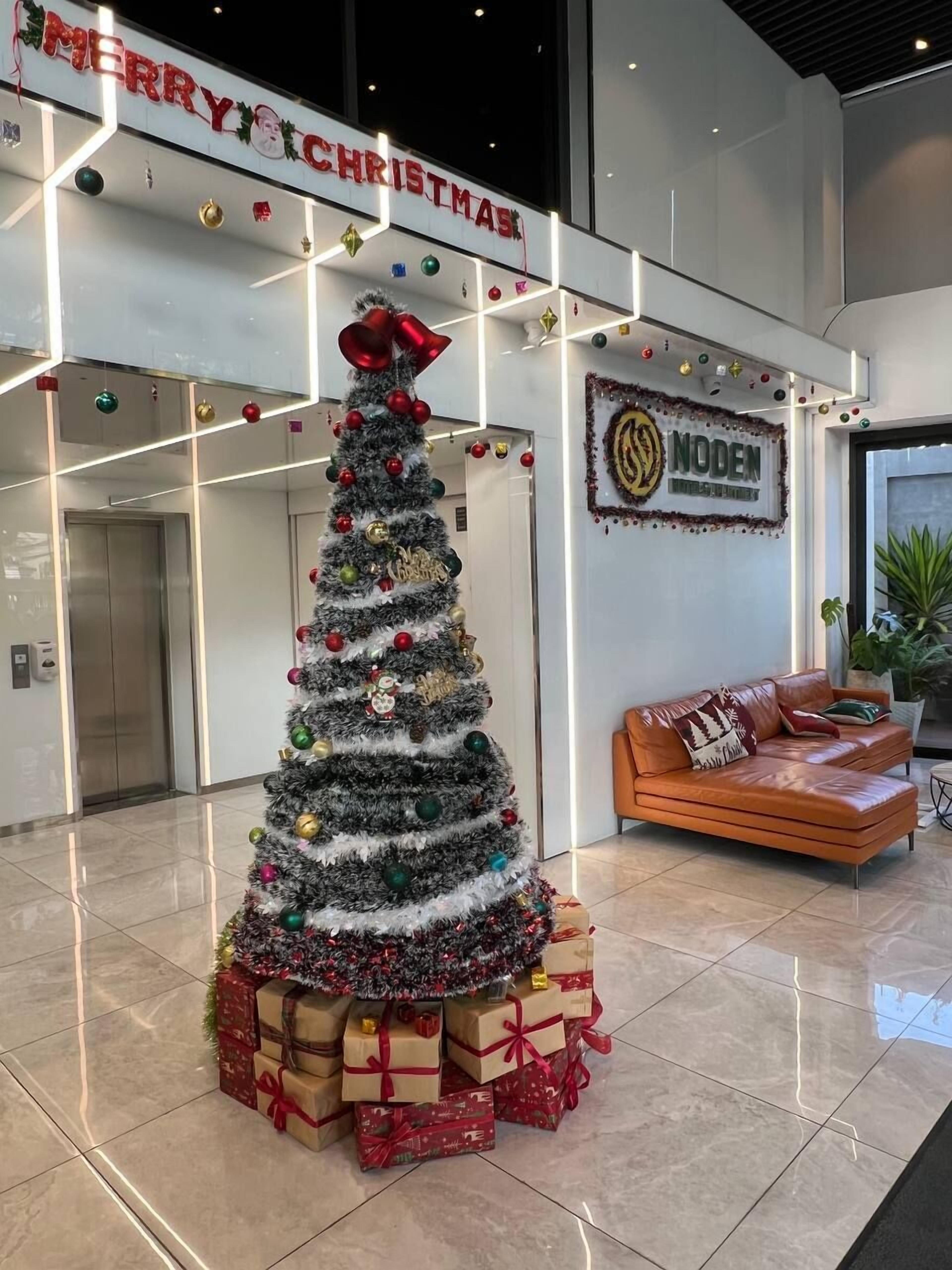 A Christmas tree in a lobby with a 'MERRY CHRISTMAS' sign above, a 'NODEN' sign on the wall, and a leather sofa with a plant.