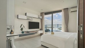 Habitación de hotel moderna con una cama grande, un televisor de pantalla plana montado en la pared, un pequeño escritorio con una planta y vistas a un paisaje urbano a través de un ventanal amplio.