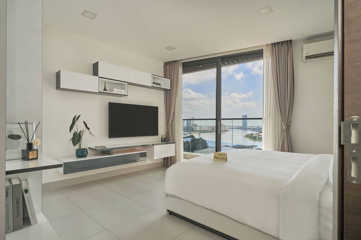 A modern hotel room with a large bed, a flat-screen TV mounted on the wall, a small desk with a plant, and a view of a cityscape through a large window.