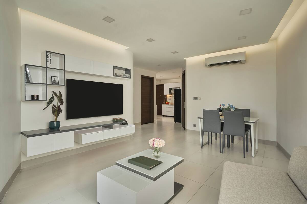 A modern living room with a flat-screen TV, white cabinetry, a dining area with chairs, and a small table with a vase of flowers.