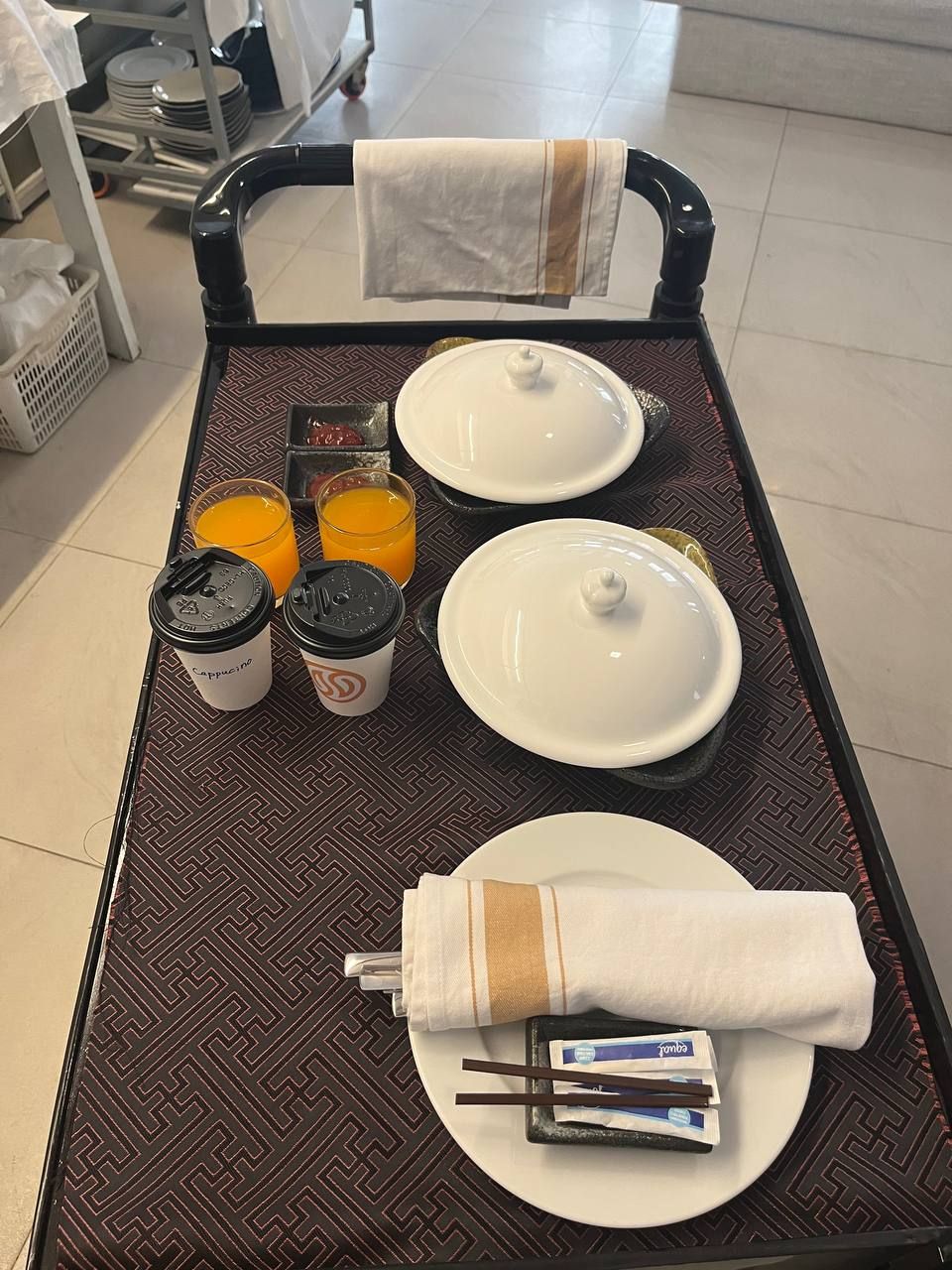 Ein Tablett mit Frühstücksgegenständen, darunter zwei weiße Schüsseln, zwei Kaffeetassen, ein Glas Orangensaft und ein gefaltetes Servietttuch mit Stäbchen und einem Instantnudelpaket.
