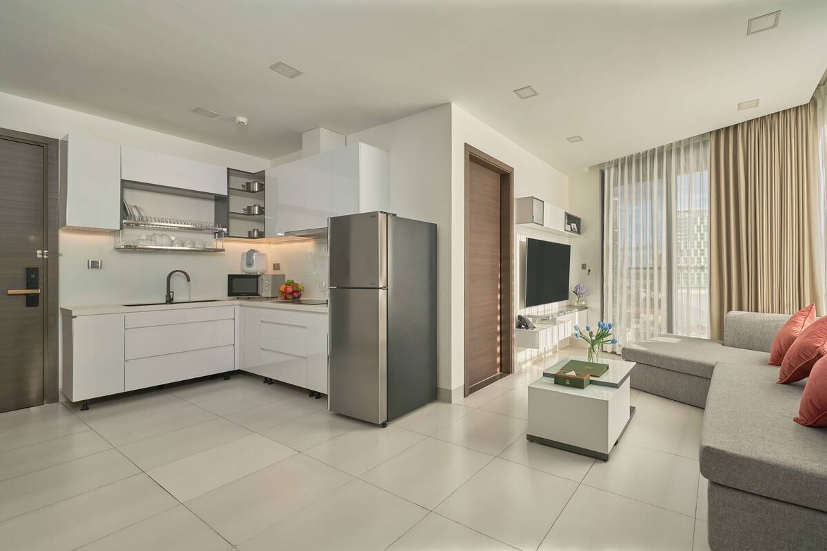 A modern kitchen with white cabinets and stainless steel appliances, a dining area with a grey sofa and red cushions, and a large window with sheer curtains.