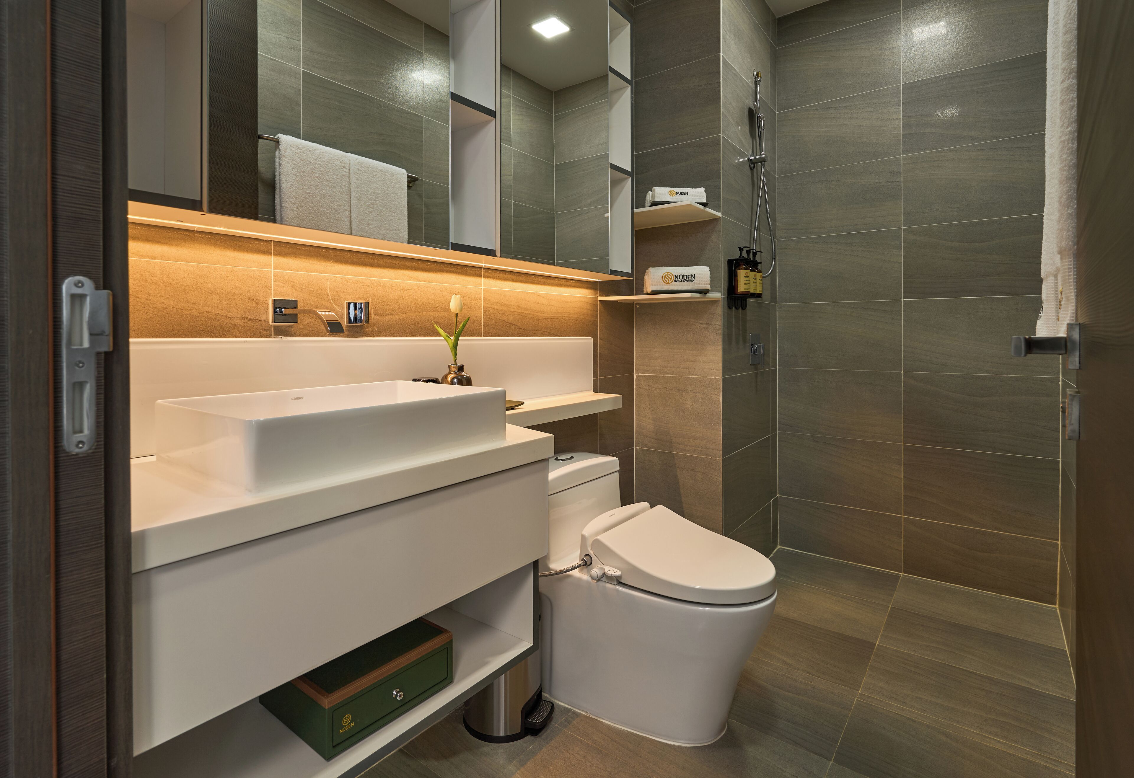 A modern bathroom with a white sink, toilet, and shower. There is a towel rack above the sink and a shelf with toiletries.