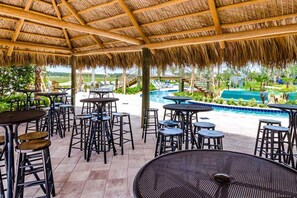 An outdoor seating area with tables and stools, a pool, and a thatched roof structure.