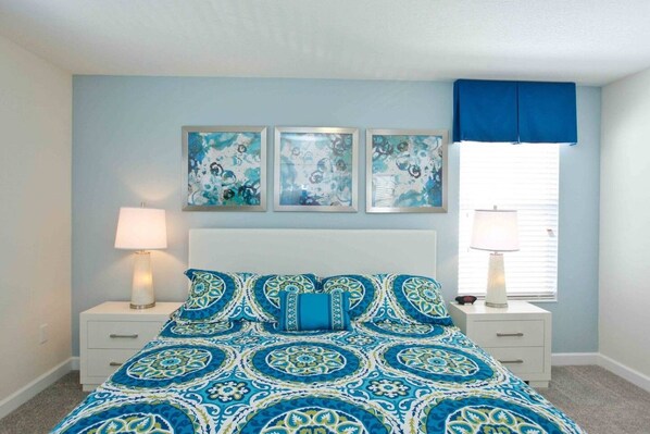 A bedroom with a patterned bedspread, two bedside lamps, a window with blue curtains, and three wall-mounted artworks.