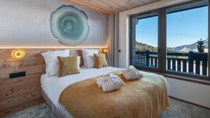 Un dormitorio con techo de madera, una cama grande con ropa blanca, una manta color arena y vistas a montañas a través de una ventana.