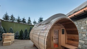 Una sauna de madera con bancos en el interior, ubicada en un muelle de madera con un muro de piedra y una valla verde al fondo.