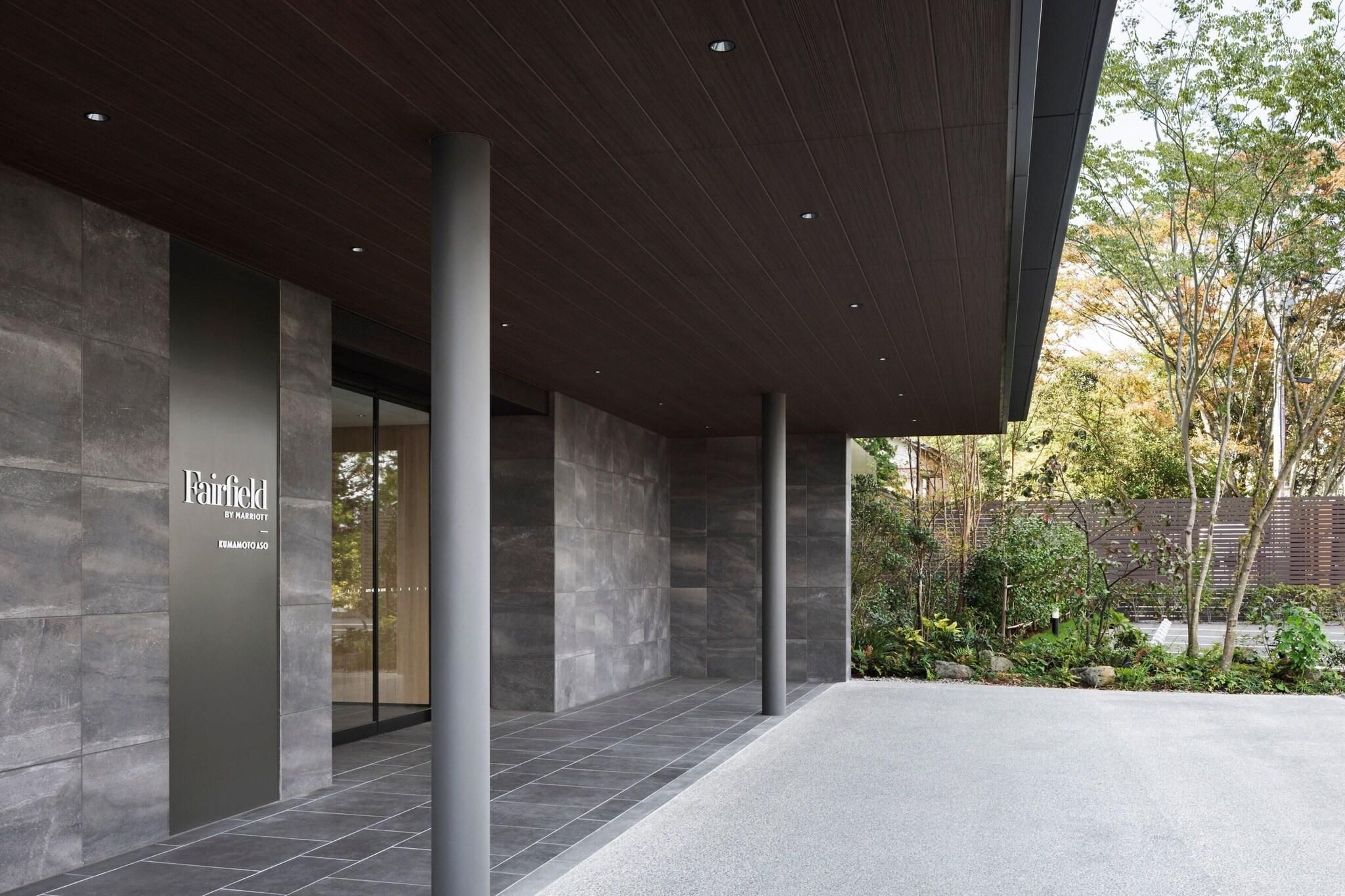 A modern building entrance with a stone facade, a sign for Fairfield by Marriott, and a landscaped garden area.