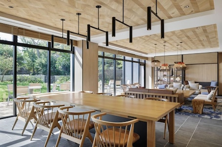 A spacious dining area with a long wooden table and chairs, large windows offering a view of the outdoors, and a modern ceiling design.