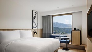 A modern hotel room with a large bed, a small table, a sofa, a TV, and a view of mountains through a large window. - Fairfield By Marriott Kumamoto Aso (Aso)