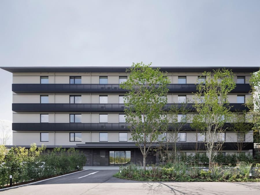 A multi-story building with a flat facade, balconies, and a parking area with trees.