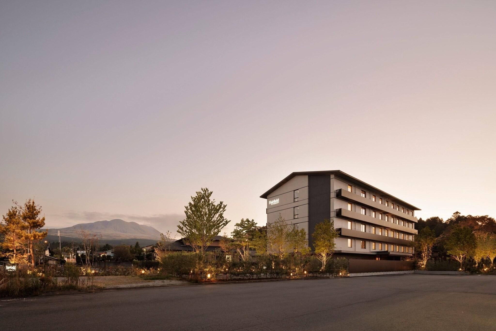 A modern hotel building with multiple floors and a flat roof, surrounded by trees and a parking lot.