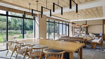 A spacious dining area with a long wooden table and chairs, large windows offering a view of the outdoors, and a modern ceiling design with hanging pendant lights.