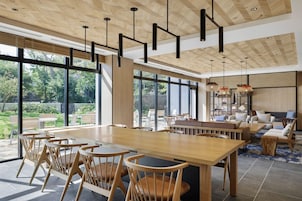 A spacious dining area with a long wooden table and chairs, large windows offering a view of the outdoors, and a modern ceiling design with hanging pendant lights.