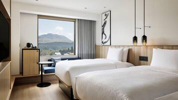 A hotel room with two beds, a small table, a TV, and a large window with a mountain view.