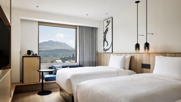 A hotel room with two beds, a small table, a TV, and a large window with a mountain view. - Fairfield By Marriott Kumamoto Aso (Aso)