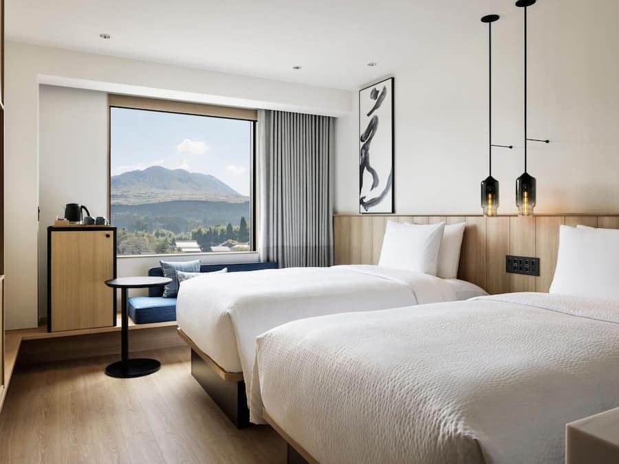 A hotel room with two beds, a small table, a TV, and a large window with a mountain view.