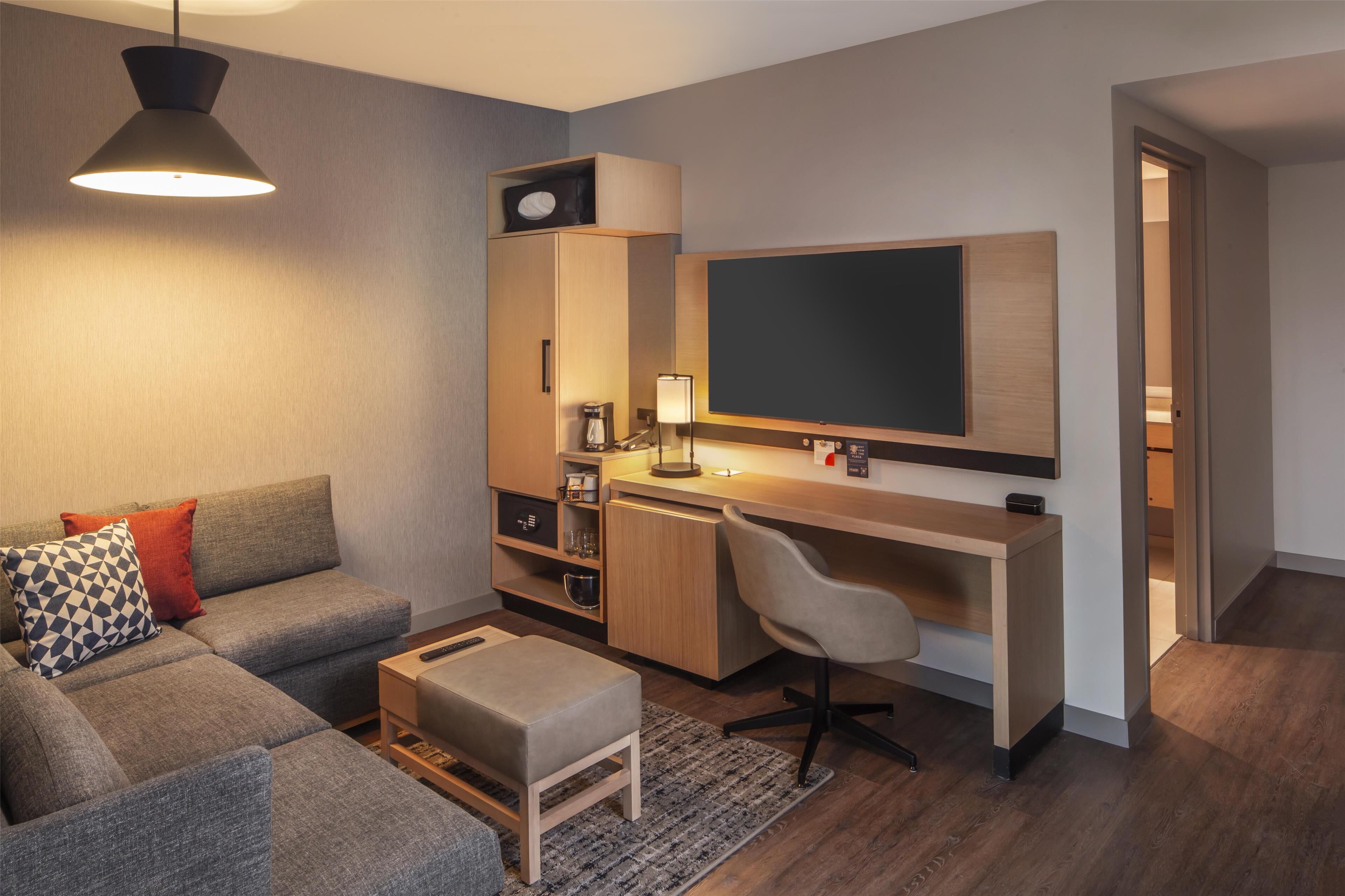 A modern living room with a grey sofa, a small ottoman, a desk with a chair, a flat-screen TV, and a coffee machine.