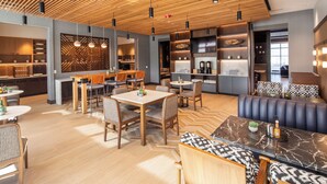 A modern hotel lobby with a wooden ceiling, a dining area with round tables and chairs, a seating area with a sofa, and a kitchen area with appliances. - Hyatt Place Reno (Reno)