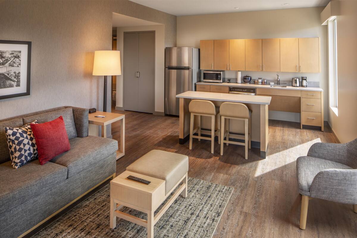 A modern living room with a grey sofa, a small table, and a coffee table. There is a kitchen area with wooden cabinets, a refrigerator, and a microwave.