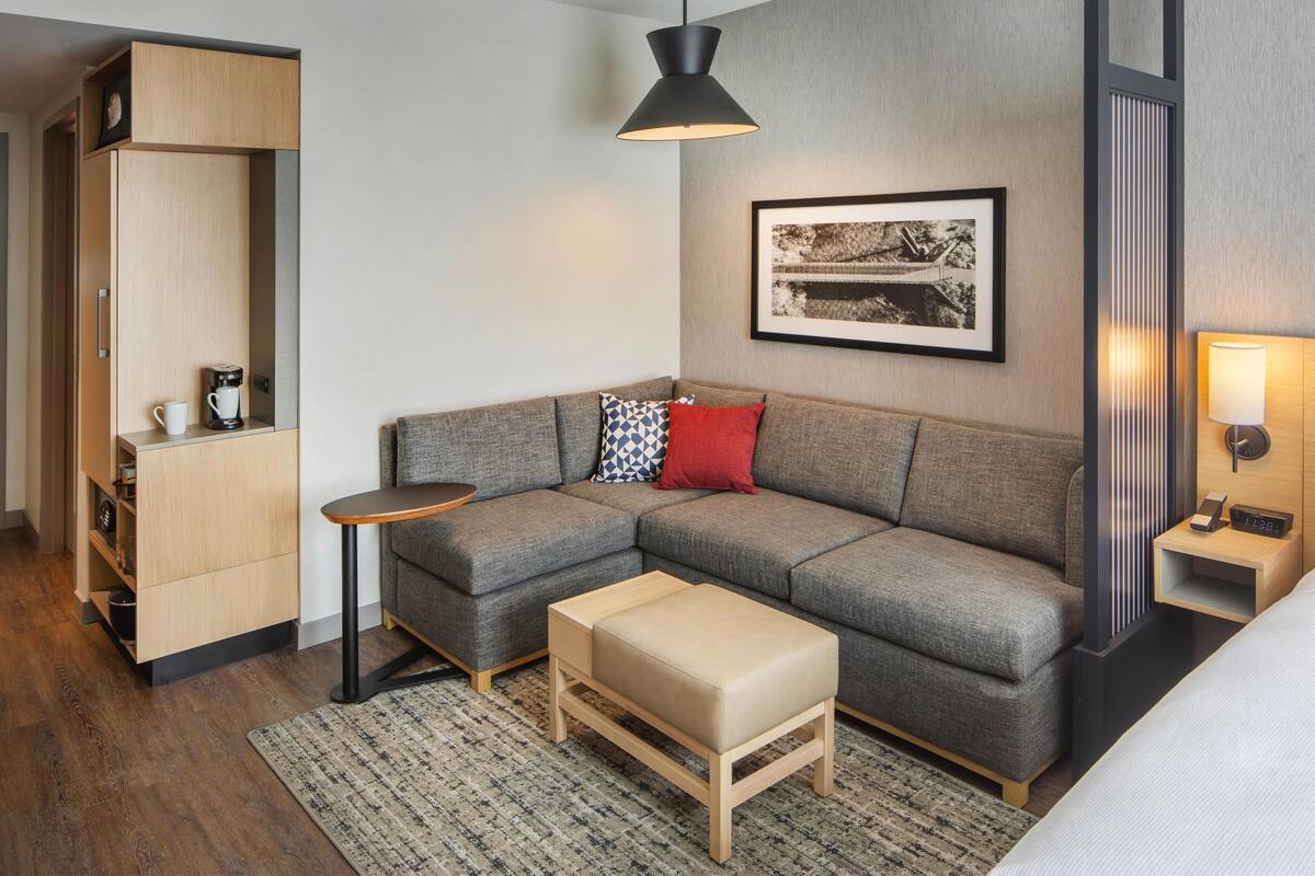 A modern living room with a sectional sofa, a small wooden table, a bench, a coffee machine, and a framed picture on the wall.