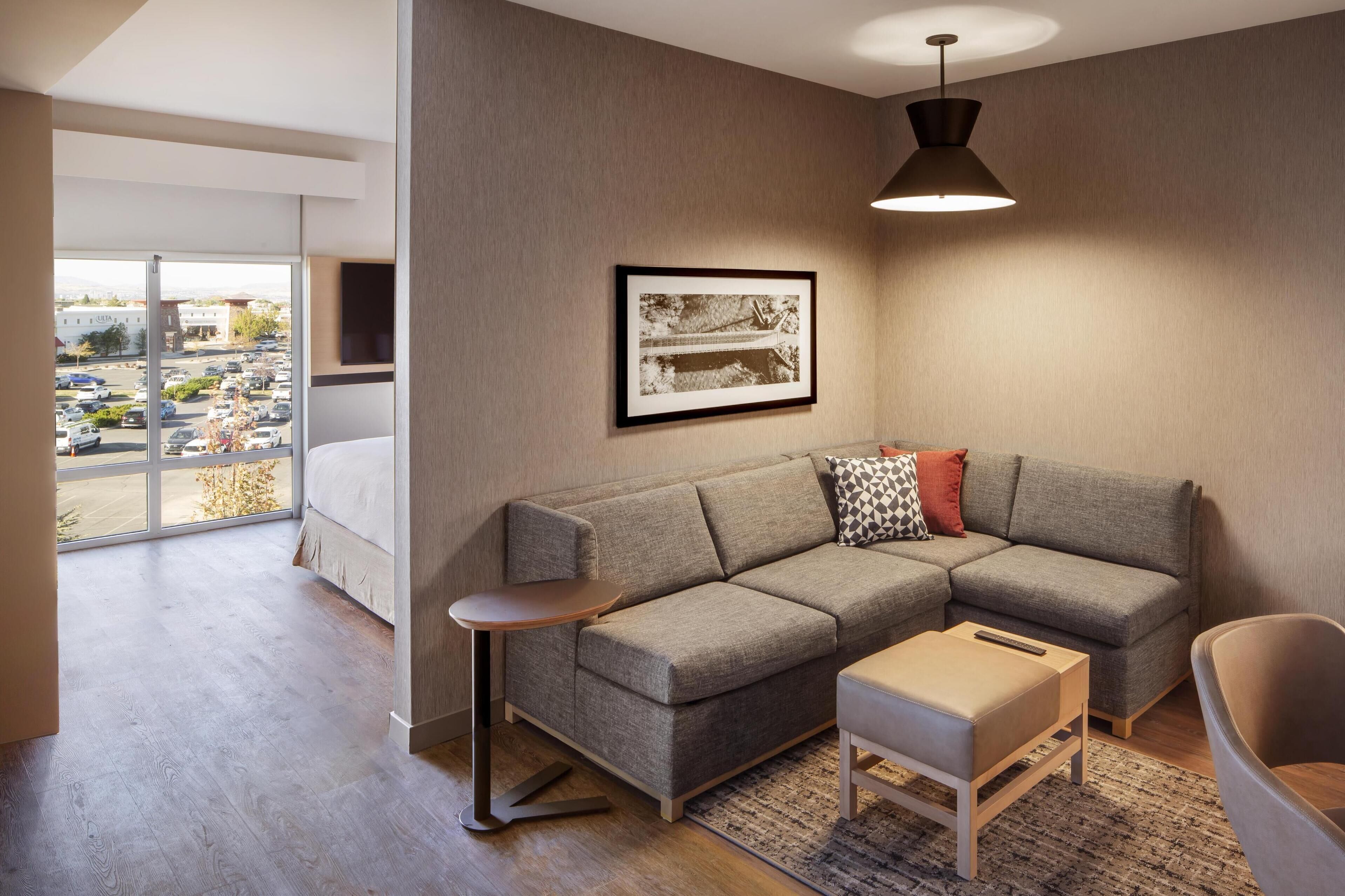 Un salon moderne avec un canapé d’angle gris, une petite table d’appoint en bois, un seul pouf et une suspension noire. Un tableau encadré est accroché au mur, et une grande fenêtre offre une vue sur un parking et des immeubles.