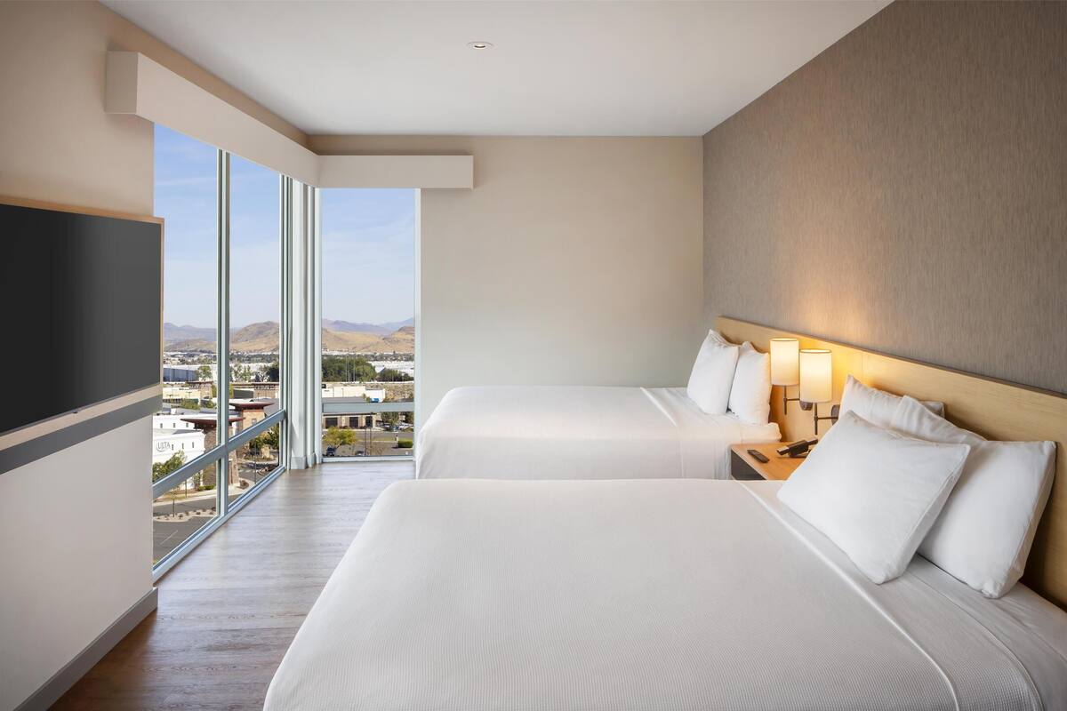 A hotel room with two beds, a TV, and a large window offering a city view.