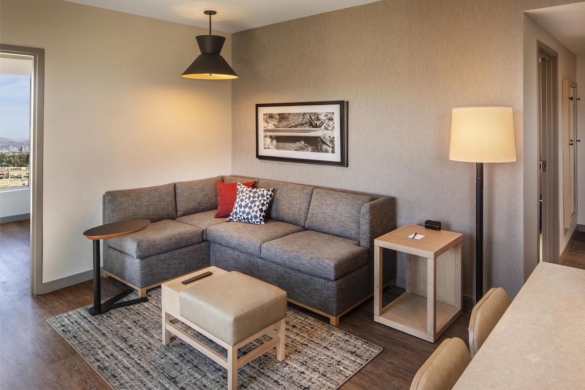 Ein modernes Wohnzimmer mit einem grauen Ecksofa, einem kleinen Holztisch, einer Stehlampe und einem gerahmten Bild an der Wand.