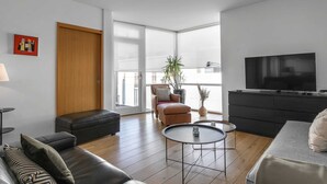 A modern living room with a sofa, ottoman, two coffee tables, a TV, a wooden door, and a balcony with a plant.