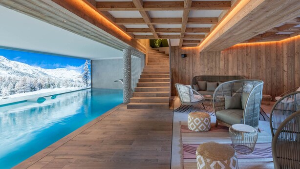 Un área de piscina interior moderna con vigas de madera, vistas a montañas nevadas y una zona de estar acogedora con cojines estampados.