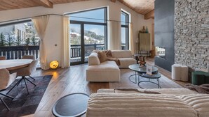 Amplia sala de estar con techo alto, vigas de madera, pared de piedra, un ventanal con vista a un paisaje nevado, un sofá, una mesa de centro y un comedor con mesa y sillas.