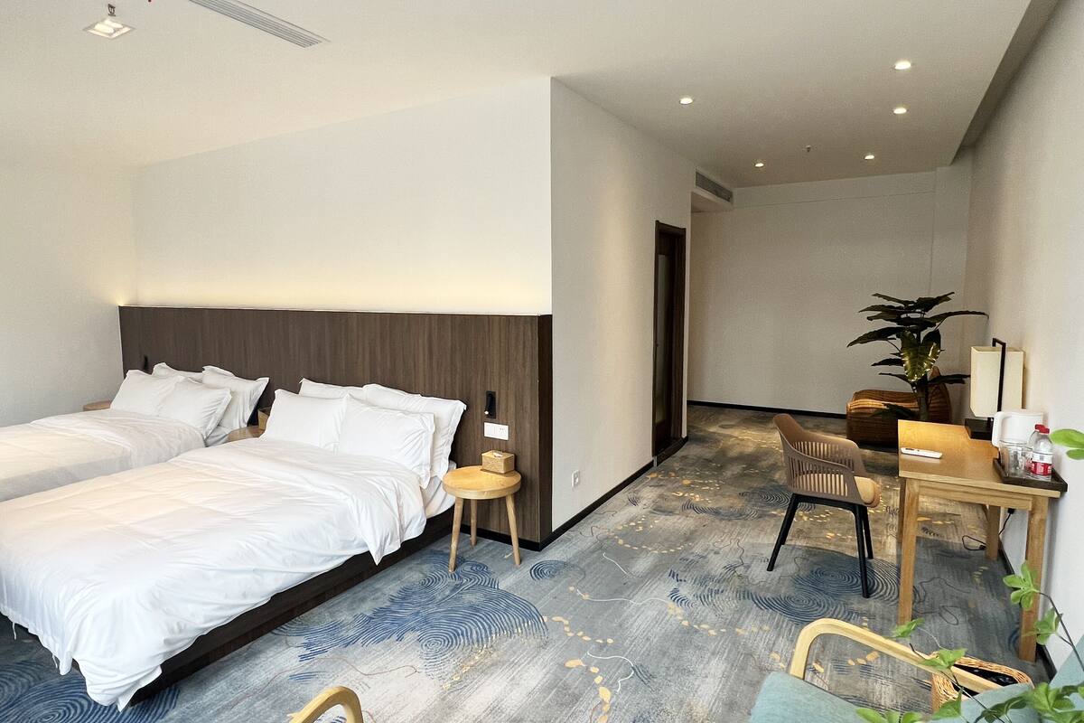 A hotel room with two beds, a desk, a chair, a small table, and a plant.
