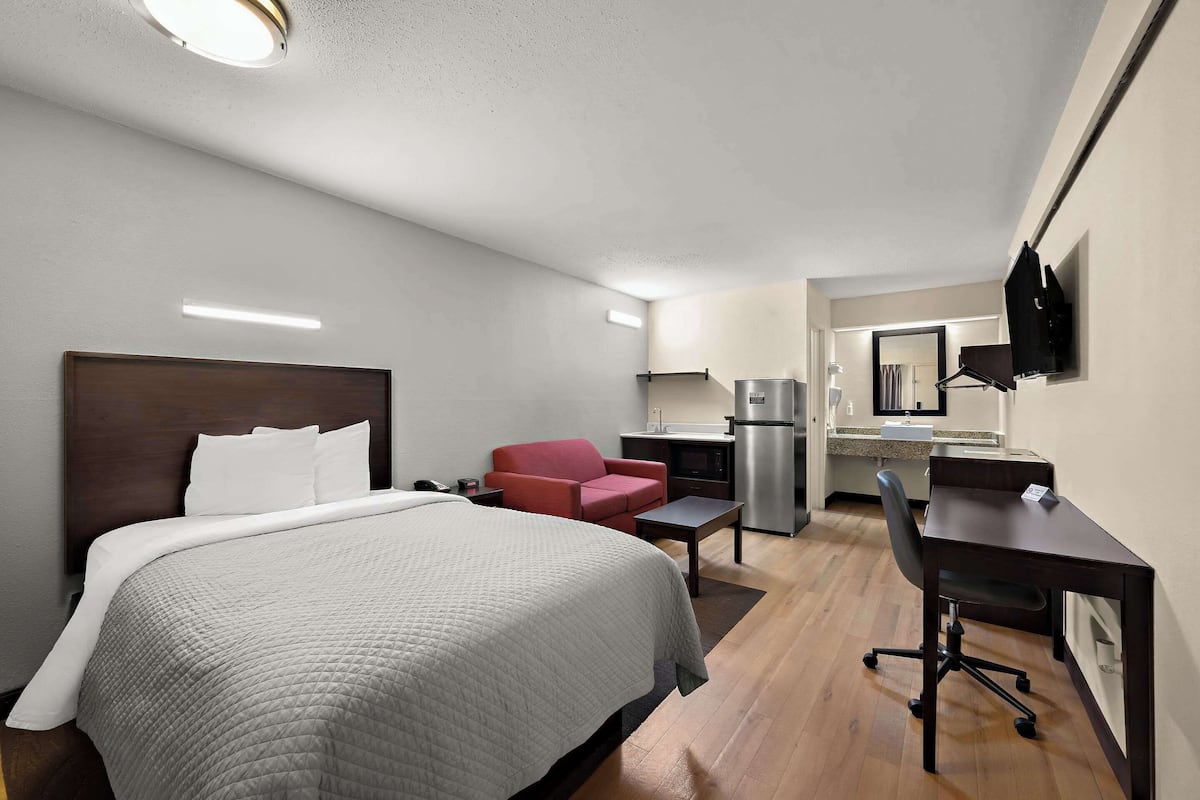 Habitación de hotel con cama, escritorio, silla, sillón rojo, kitchenette y televisor montado en la pared.